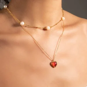 Delicate layered 18K gold necklaces on female model, heart pendant with red stone worn with pearl choker on bare neck and collarbone