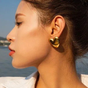 18K gold heart earrings worn on ear side profile close-up showing smooth gold metal design on model with tied-back hair outdoor waterside setting