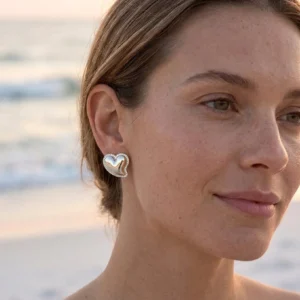 Silver-gold heart stud earrings close-up side profile on female model at coastal ocean beach setting, luxury jewelry photography
