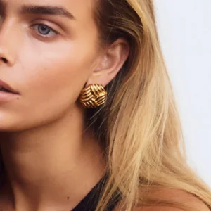 Close-up side view of woven 18K gold hexagon stud earring worn on blonde model's ear, showcasing detailed ribbed texture and geometric design