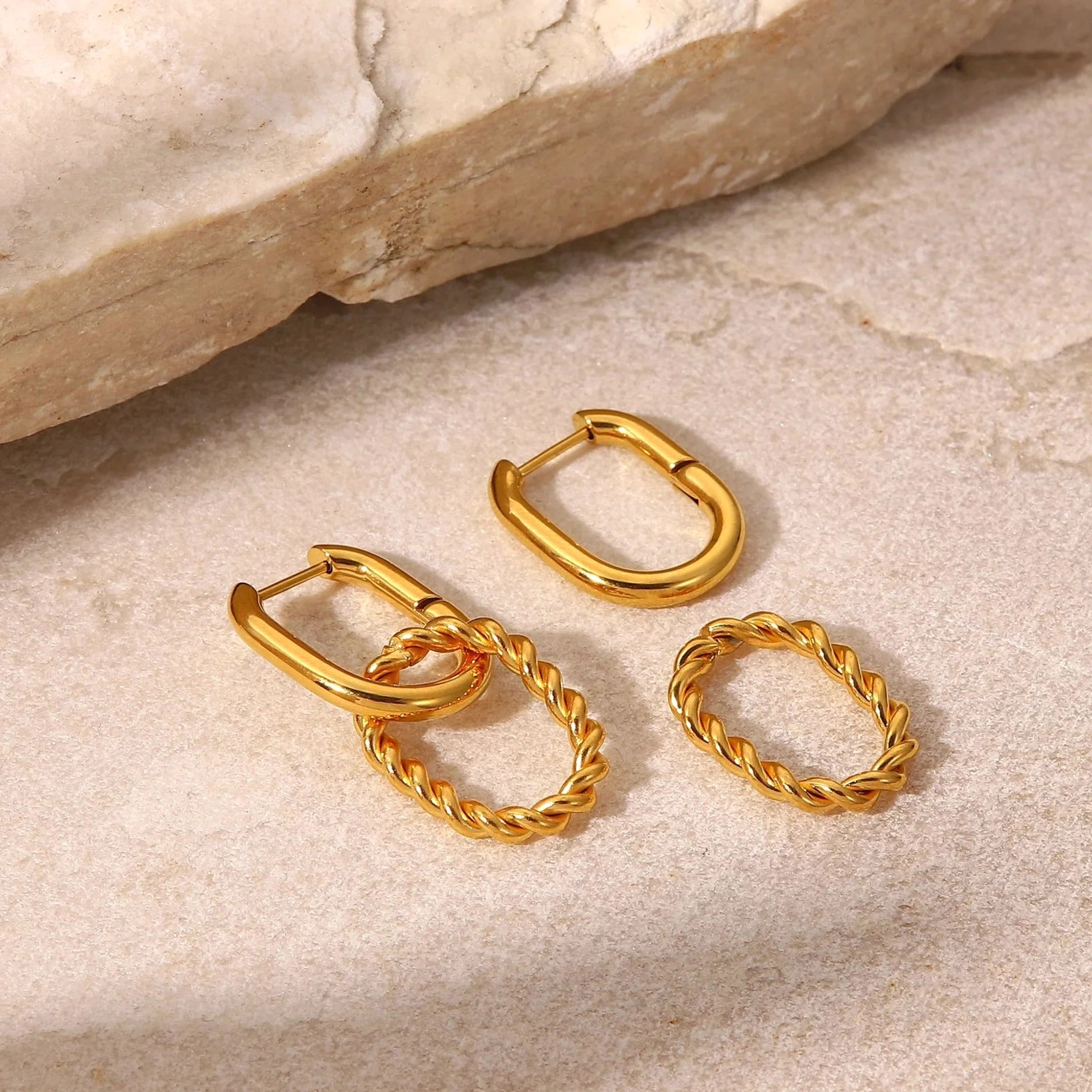 Top-down flat lay of mixed 18K gold hoop earrings on natural stone surface, showing smooth oval huggies and twisted rope texture variants