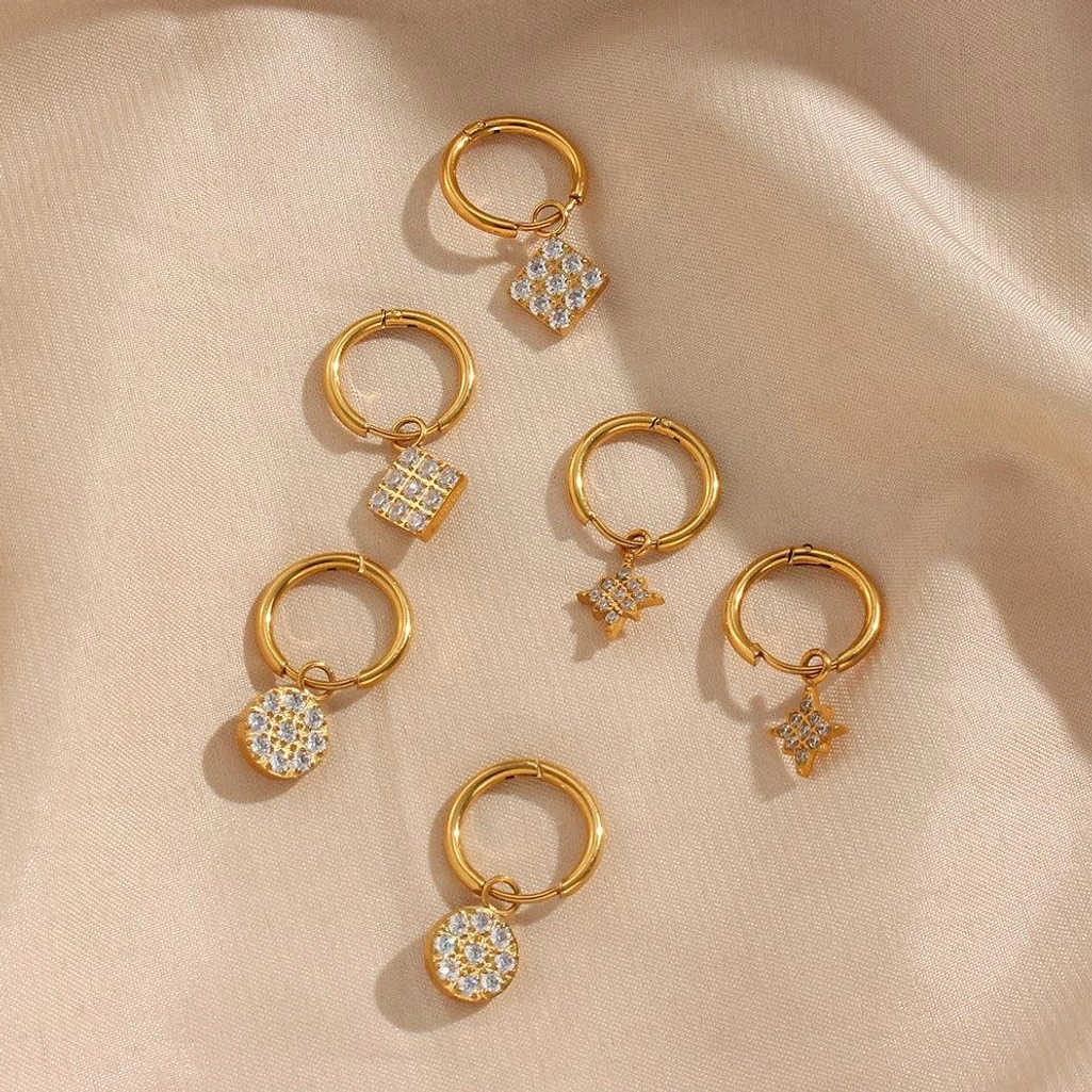 Assorted 18K gold huggie hoop earrings with multiple cubic zirconia charm styles flat lay satin fabric