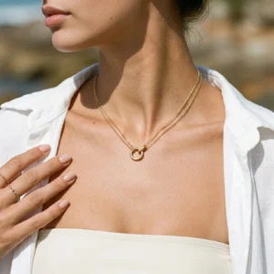 Double layered gold circle pendant necklace worn by female model in casual white outfit outdoors at seaside with shiny interlocking hoop pendant