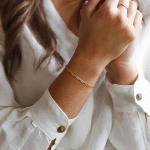 Gold paperclip chain bracelet worn on female wrist close-up, delicate thin chain design paired with white linen blouse, everyday jewelry styling