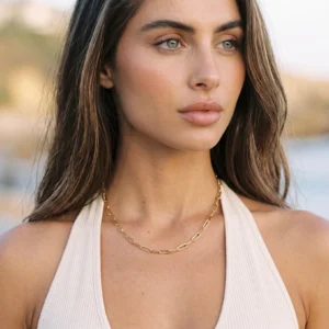 Woman wearing 18K gold paperclip chain necklace at collarbone positioned on chest paired with white halter top outdoor seaside setting