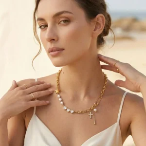 Model wearing 18K gold pearl diamond cross charm necklace with white slip top outdoors by seaside water elegant styling
