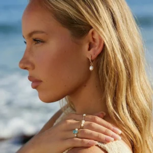 Delicate 18K gold pearl drop earrings worn by blonde woman at seaside beach, side profile close-up showcasing minimalist design