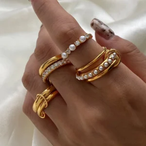 Woman wearing layered 18K gold pearl rings with rhinestone accents on manicured hand with satin backdrop