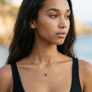 woman wearing red birthstone necklace outdoors - 18K gold pendant jewelry on coastal background