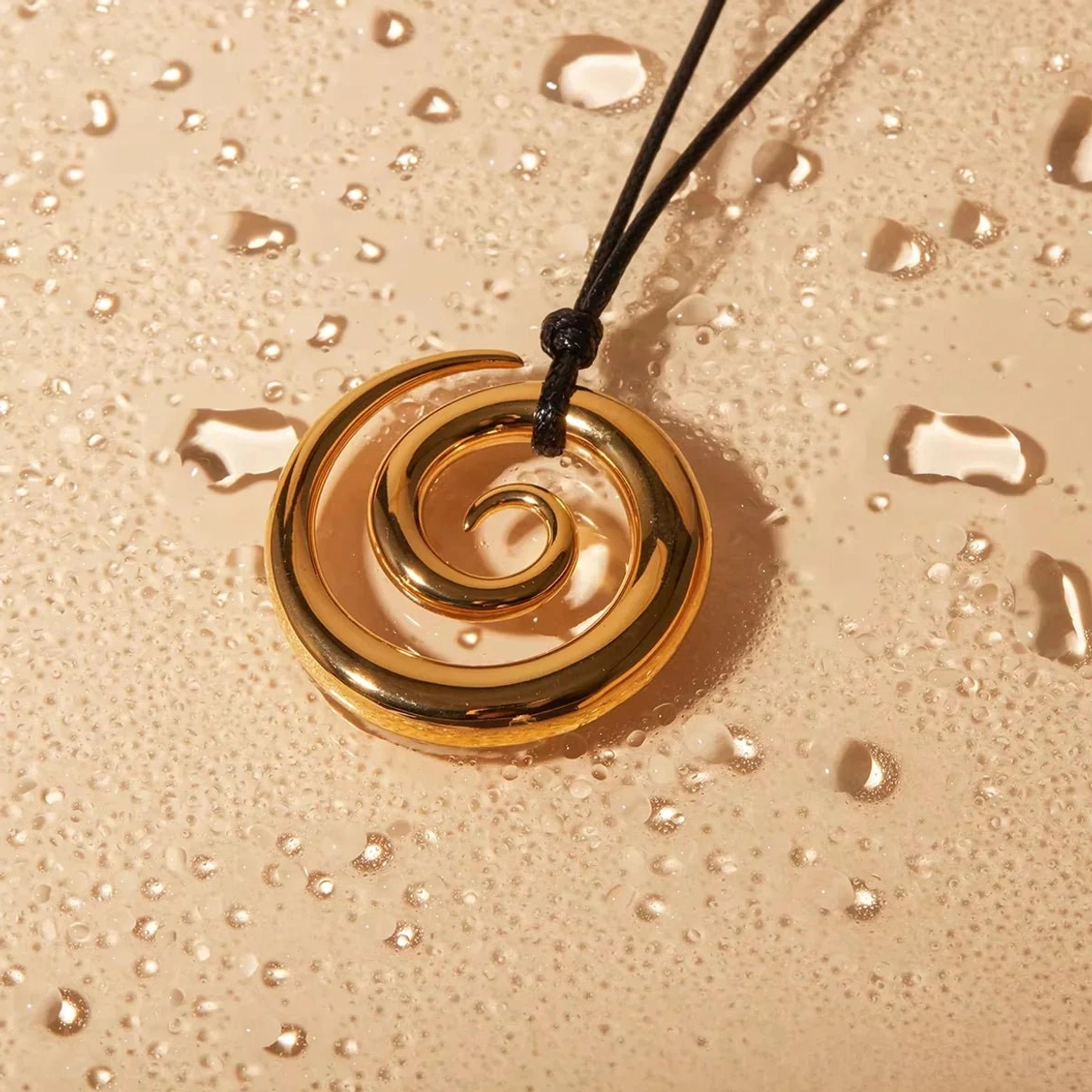 Glossy 18K gold spiral pendant with knotted black cord on water droplet textured background - water-resistant durability showcase