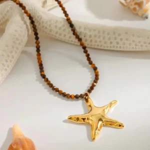 Close-up angled view of 18K gold starfish pendant with polished glossy charm and tiger-eye tone beads styled with nautical seashell and starfish props