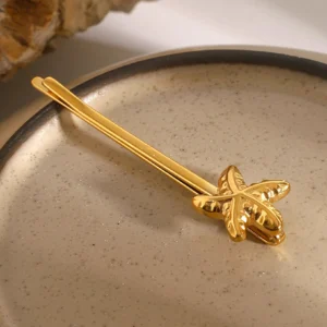 3D textured starfish bobby pin in warm gold tone, top-down close-up on ceramic dish with stone decor, premium finish