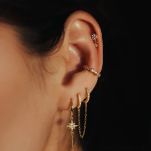 Gold piercing earring stud close-up side view on ear model wearing multiple gold jewelry pieces dark background