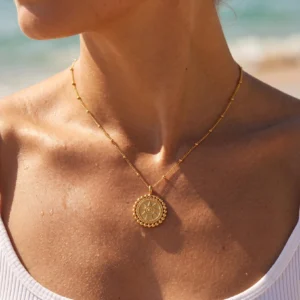 Woman wearing 18K gold sun pendant necklace on beach near ocean water, close-up of collarbone