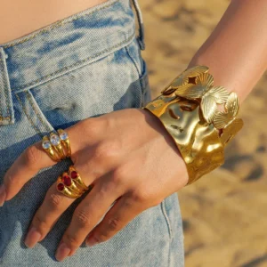 Wide gold cuff bracelet with butterfly embossed design worn with stackable gem rings on woman wrist at beach outdoor setting