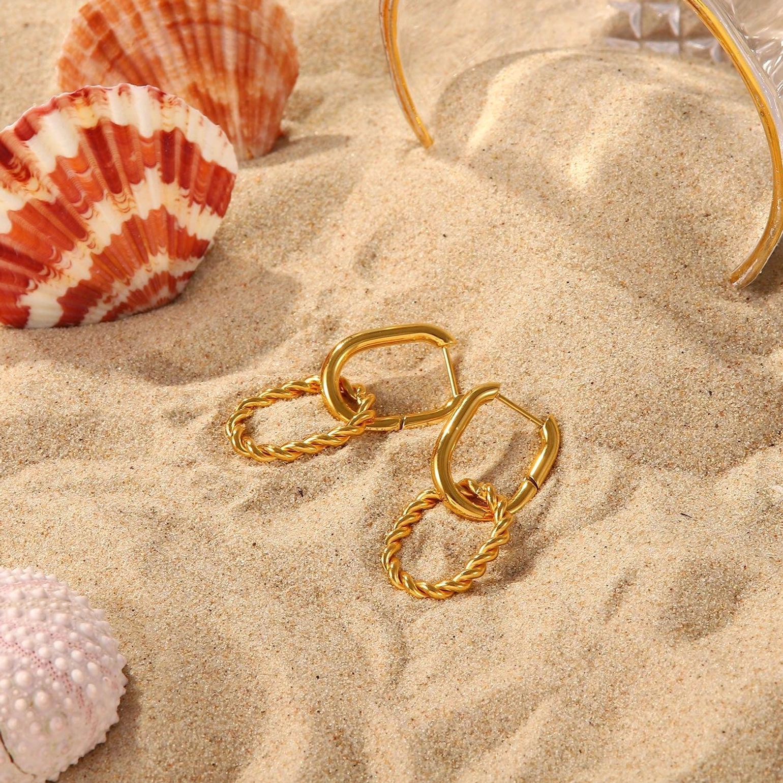 Overhead flat lay of 18K gold twisted hoop earrings on beach sand with seashell decor, demonstrating water-resistant hypoallergenic design