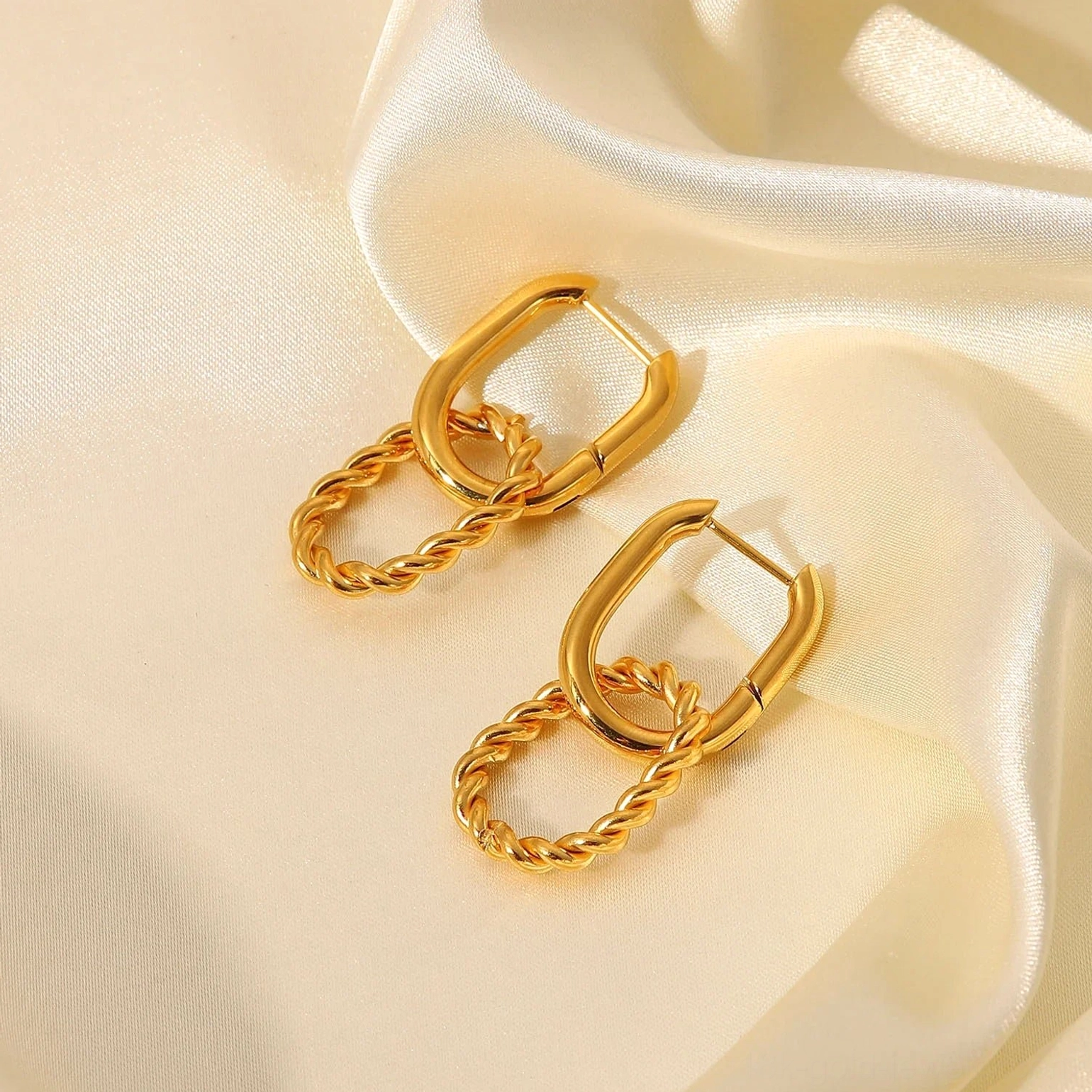 Top-down flat lay of 18K gold huggie hoop with twisted rope oval drop pendant on satin silk fabric, luxury jewelry styling