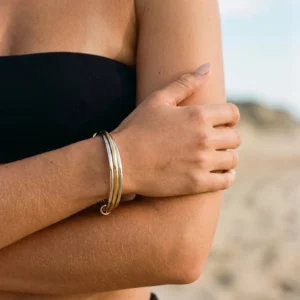Two-tone gold and silver stacked bangles worn on wrist at beach, close-up forearm detail with hinged clasp