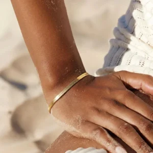 Black model wearing slim 18K gold herringbone bracelet on wrist at beach with sand and ocean background lifestyle photo