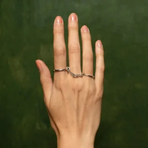 Chain dual-finger ring worn on female hand with nude manicure back-of-hand perspective dark green backdrop minimalist silver jewelry
