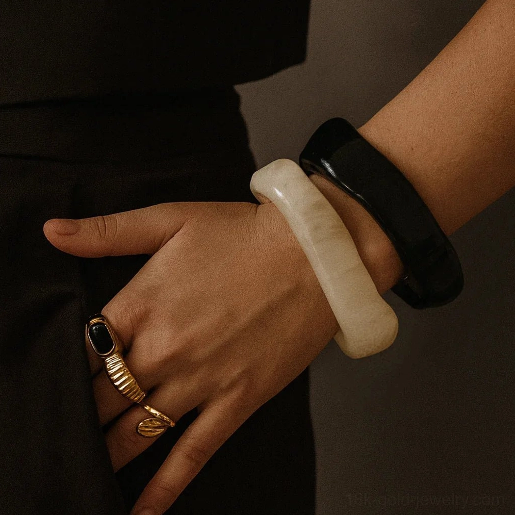 Female model wearing stacked colorful acrylic bangles with cocktail rings on wrist, featuring off-white and black rounded square bangles against dark neutral background