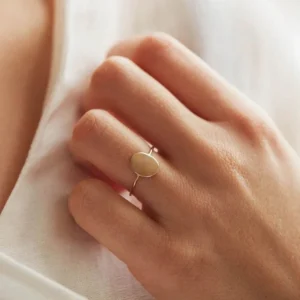 Eleanor gold oval signet ring close-up worn on female middle finger against white fabric