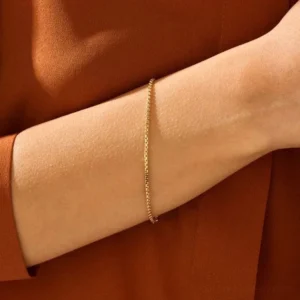 thin gold chain bracelet worn on wrist, close-up side view, model wearing burnt orange sleeve, delicate slim box chain