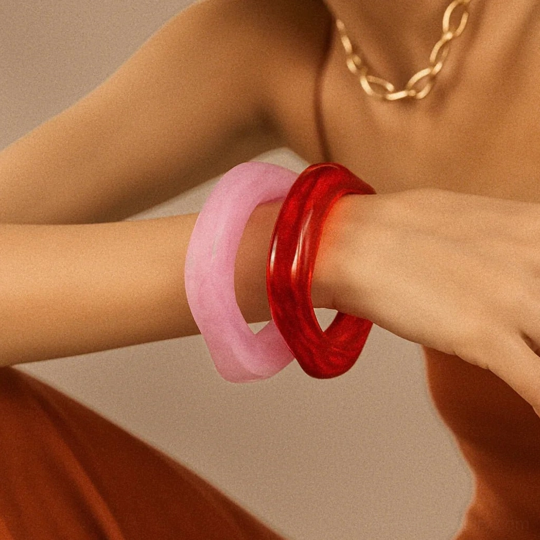 Pink opaque and bright red glossy dual acrylic bangles on female model wrist with gold chain, warm studio lighting showcasing rounded square shape