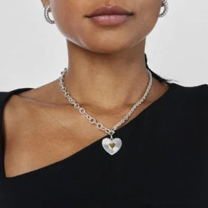 Mixed metal two-tone heart pendant necklace worn on female model with black off-shoulder top, pendant rests at collarbone, lifestyle styling with small hoop earrings